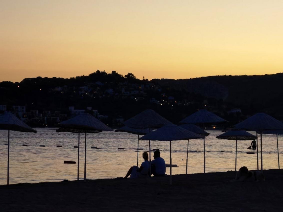 A lively scene of Bodrum’s beachfront resorts with vibrant nightlife, beach clubs, and the historic Bodrum Castle under a sunset sky.